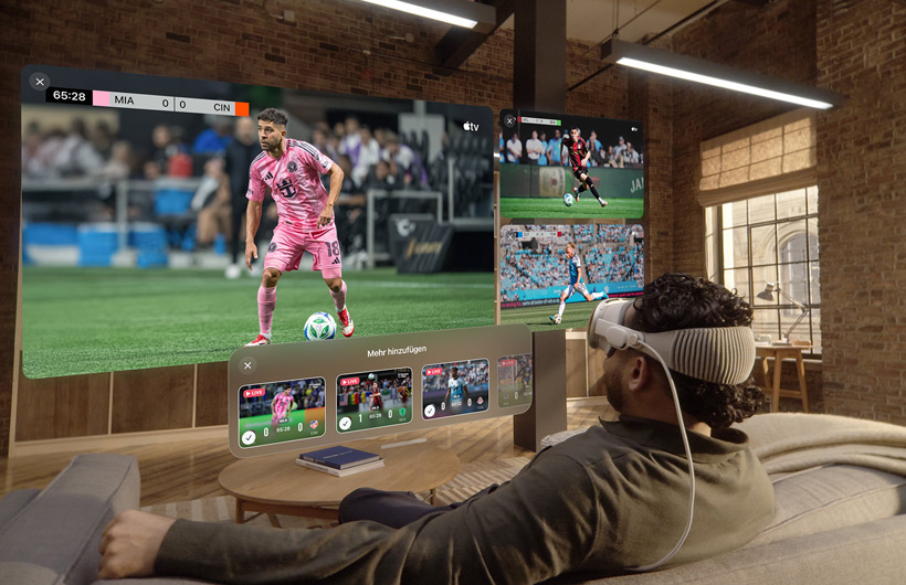 Eine Person mit einer Apple Vision Pro sitzt auf einem Sofa in einem Wohnzimmer. Vor ihr wird ein großer Videobildschirm neben zwei kleineren Bildschirmen gezeigt, die jeweils ein anderes Sportereignis zeigen. Unter dem großen Bildschirm befindet sich ein Auswahlfenster mit dem Text „Mehr hinzufügen“