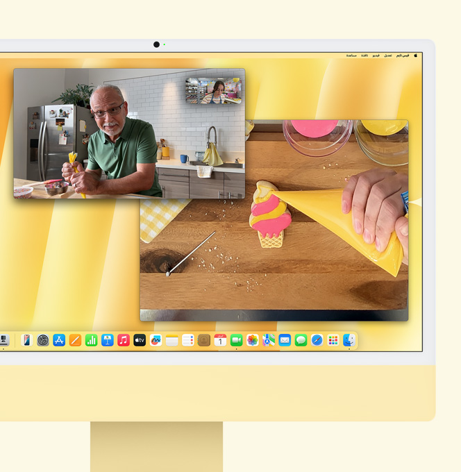 iMac باللون الأصفر. عرض توضيحي لميزة العرض الرأسي. شاشة تعرض صانعة حلويات تشارك في مكالمة فيديو مع شخص آخر. وتعرض إحدى الشاشتين صانعة الحلويات ووجه الرجل، بينما تعرض الشاشة الأخرى يدي صانعة الحلويات وهي تزيّن قطعة بسكويت