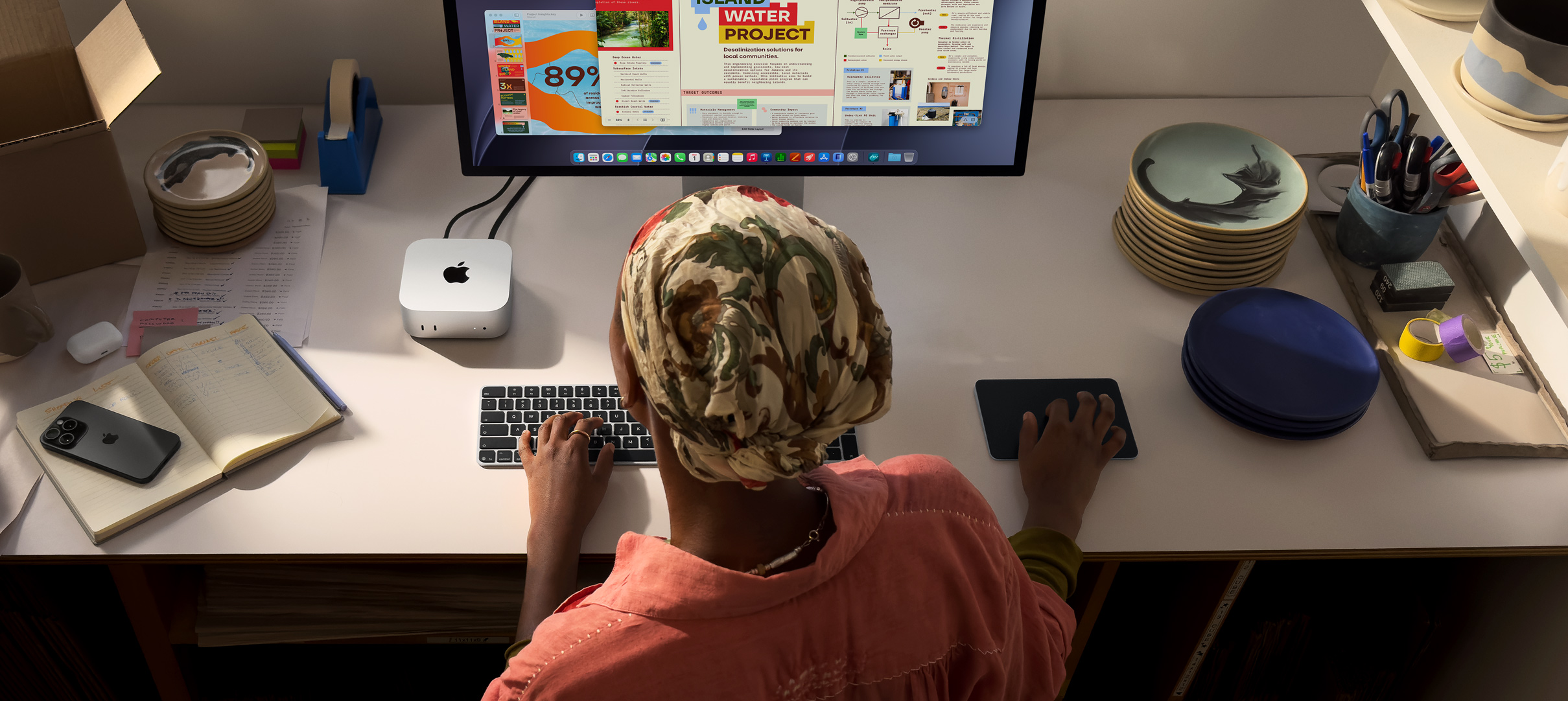 سيدة تطبع وهي جالسة إلى مكتب مع جهاز Mac mini، شاشة 
Studio Display متصلة، لوحة مفاتيح ماجيك، لوحة تعقب ماجيك، أغراض متنوعة
