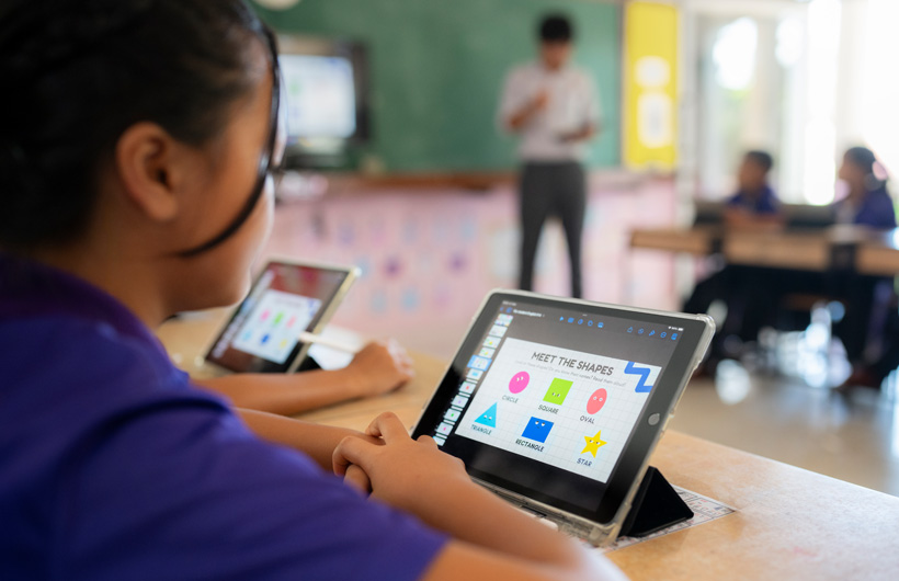 Close-up of a student at their desk, using an iPad. On screen, there are colourful shapes displayed with the title, 