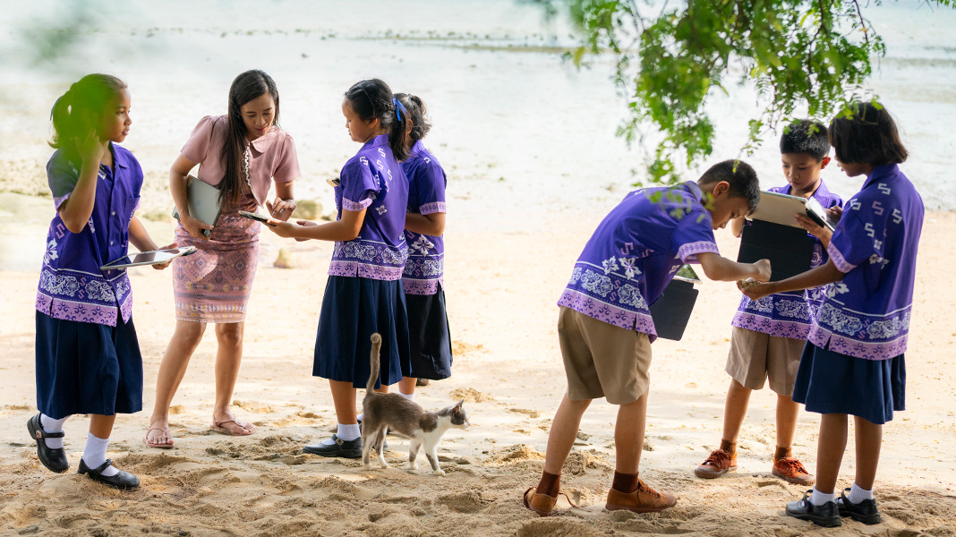 Six elementary students in colourful purple uniforms stand on a sandy beach holding iPads. A teacher is assisting one student on an iPad, and there is a cat walking amongst them.