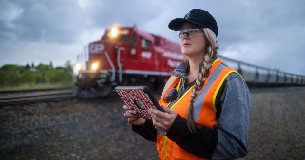 Eine Bahnbedienstete hält ein iPad in der Hand und schaut auf die Gleise, während ein Zug vorbeifährt