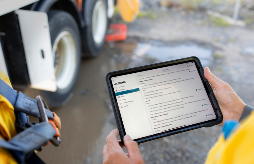 Handen houden een iPad vast met de HazChat-app geopend die taakdetails, planning, gevaren en controles weergeeft, terwijl een persoon op een werkplek bij een modderpoel staat