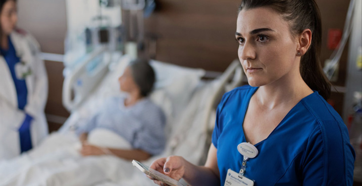 Eine Pflegekraft schreibt auf ihrem iPhone eine Textnachricht, während eine andere Medizinerin mit einer Patientin spricht.
